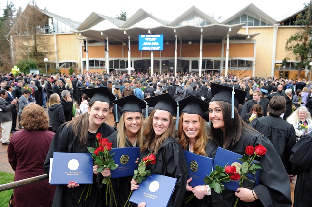 Five graduates in cap and gown gather in front of the Rec Center, holding diplomas and roses.